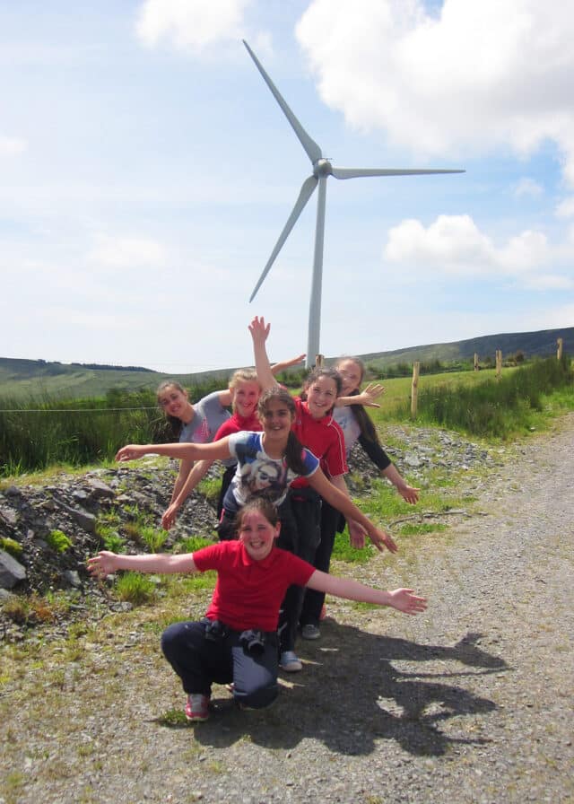 Young school pupils make windmill shapes in front of wind turbine