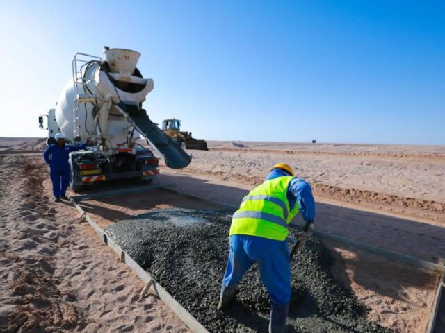 Construction worker with his back to the camera spreading cement that is being poured by a cement truck.