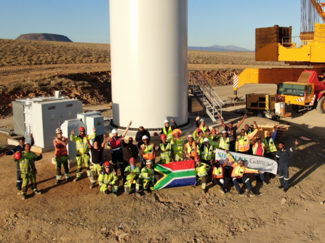 local construction workforce - about 25, waving to the camera from the base of a wind turbine