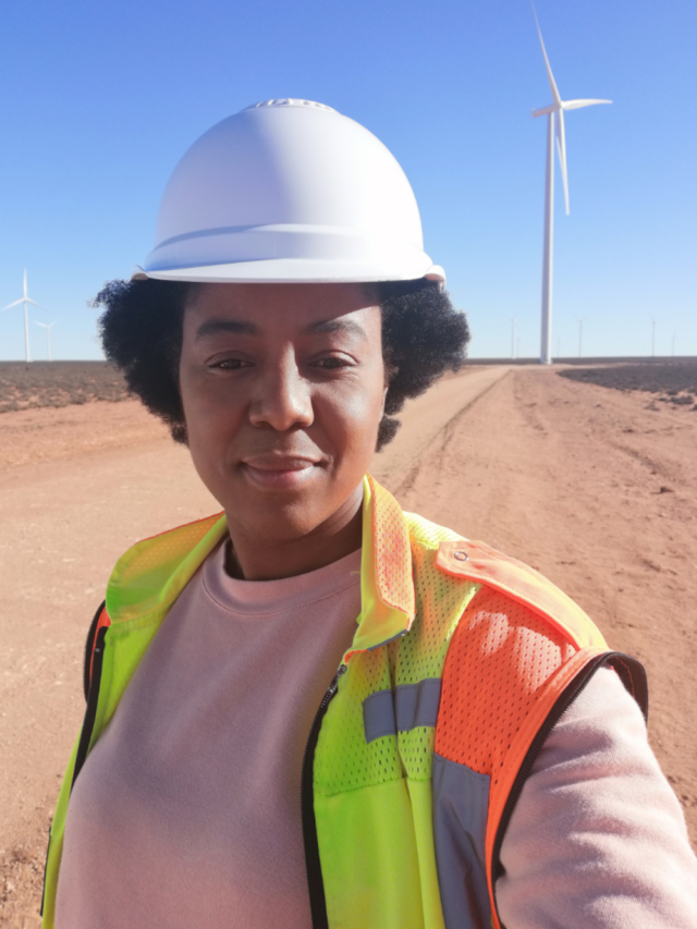 A picture of of Miliswa with a high vis and hard hat on in front of a wind turbine.