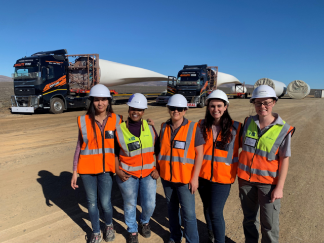 5 women with hard hats and high vis on standing in front of long haul trucks with wind turbines on the trailers.