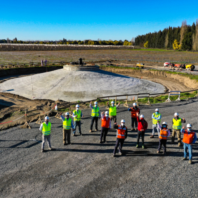 Waving team members in hi-visibility jackets standing at the base of a wind turbine's foundation
