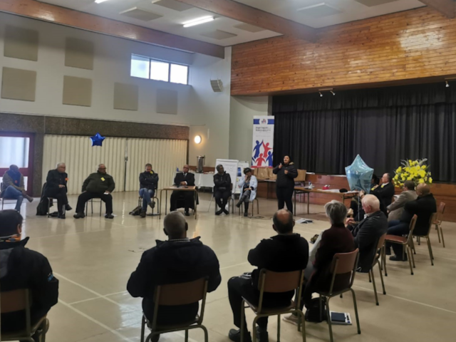 Group of people sitting on chairs in a circle in a hall, listening to someone standing speaking.